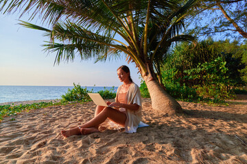 Young contented joyful happy girl digital nomad freelancer sitting on a sand under a palm tree on...
