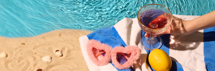 Sandy beach setup with pink fuzzy sunglasses, lemon, and glass of drink. Blue towel adds color against serene pool waters, creating refreshing summer aesthetic. Concept of summer, relax, tourism. Ad