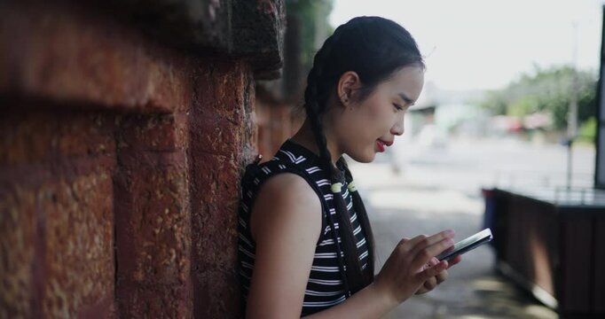 Handheld side view shot, Attractive Asian woman leaning against a wall on a sidewalk enjoying the city view while using a map app to find directions on mobile phone.