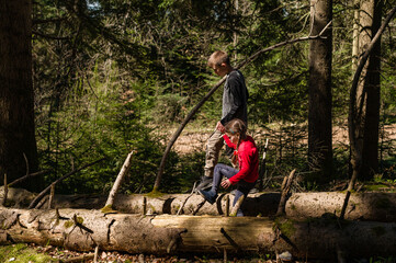Fototapeta premium Two children, a boy and a girl, navigate a fallen log in a lush forest. Sunlight filters through the trees, creating a magical atmosphere. A scene of sibling companionship and outdoor exploration.