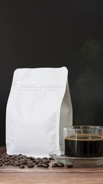 4K vertical. Roasted coffee beans in white paper bag with zipper on wooden table, blank mockup coffee bag template for product design advertising, black coffee hot stream in coffee cup on set