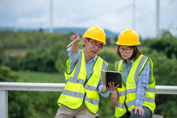 Two workers wearing safety vests and hard hats are looking at a tablet