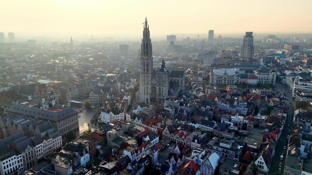 Under a hazy morning sky, a panoramic aerial view reveals Antwerp's historic heart, where the towering spire of the Cathedral of Our Lady dominates a tapestry of red-tiled roofs and winding streets