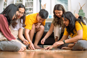 A group of Asian friends gathers indoors, enthusiastically collaborating on a puzzle game. They sit on the floor, fully engaged, with a cozy background featuring shelves, decor, and a modern interior.
