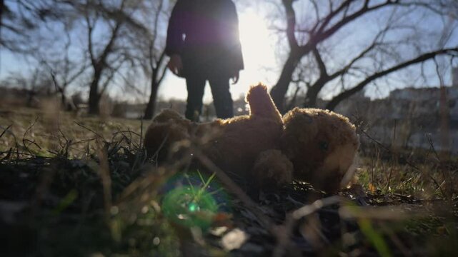 A joyful little girl found her lost soft toy. A child approaches an abandoned teddy bear and picks it up from the ground on a sunny day in the park.