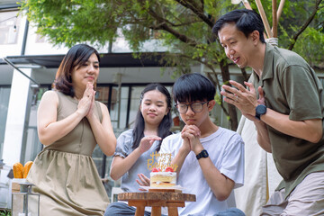 An Asian family gathered outdoors, celebrating a birthday. A young boy is making a wish with his hands clasped, as others clap and smile. A small cake with candles is placed on a wooden stool
