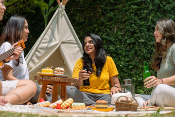 A diverse group of Asian friends enjoying a cheerful outdoor picnic with drinks and snacks. They sit on a blanket near a tipi, sharing laughs amidst a lush green garden under bright sunlight