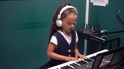 Cute singer is recording a new song for her music album in a production studio. Little girl artist musician wearing headphones playing digital keyboard electronic piano and singing a song.