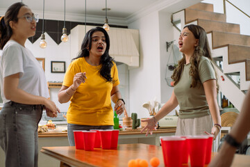 Asian friends enthusiastically playing beer pong in a lively indoor setting. Red cups and orange...