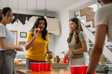 Asian friends enthusiastically playing beer pong in a lively indoor setting. Red cups and orange ping-pong balls are on the wooden table, surrounded by laughter and cheerful interaction.