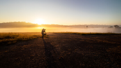 Landscape photographer takes a photo alone in the grass, in the morning, with mist and the sun in the background.