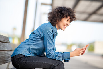Side-view portrait of a curly-haired smiling woman using a phone while sitting outdoors