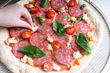 A child's hand places a basil leaf on a raw pepperoni pizza with tomatoes and cheese.