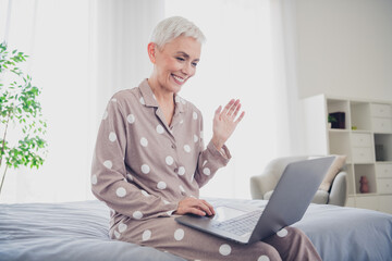 Cheerful senior woman in polka dot pajamas waving while using laptop in cozy bedroom