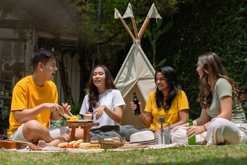 A diverse group of Asian friends enjoying a cheerful outdoor picnic with drinks and snacks. They...
