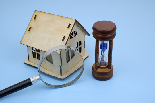 Wooden house model under magnifying glass and hourglass on blue background.