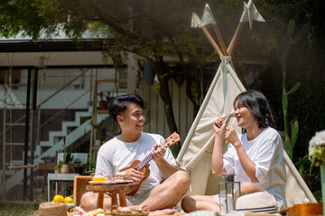 Asian couple enjoying a lively picnic in a serene garden setting. The man plays a ukulele while the...
