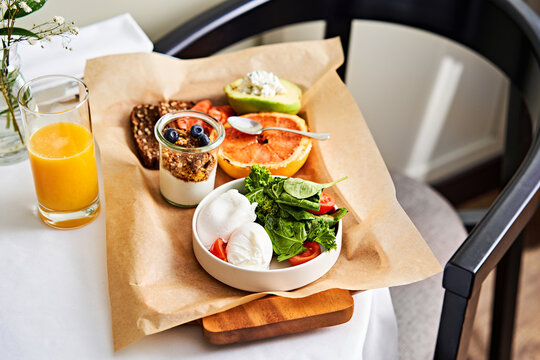 A colorful breakfast is presented on a wooden tray at a cafe table. The meal includes fresh fruits, creamy yogurt with berries, a salad of greens and tomatoes, alongside refreshing juice