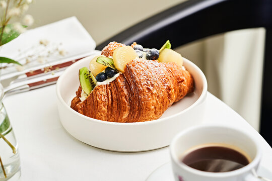 A delicious croissant is elegantly arranged with fresh fruits on a white plate beside a steaming cup of coffee, creating a warm breakfast setting