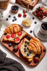 Delicious breakfast spread featuring buttery croissants topped with fresh strawberries and blueberries, served on a wooden board with butter, coffee, and fruit, creating a cozy