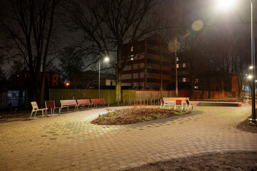 Obraz premium A small urban park at night with a curved pathway, red and white benches, sparse vegetation, and multi story buildings in the background.