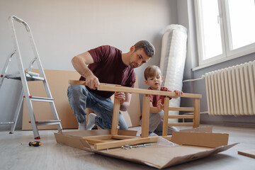 A father and his young son assembling wooden furniture at home, promoting teamwork, skill-building,...