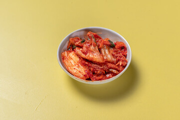 A small white bowl filled with traditional Korean kimchi, placed on a vibrant yellow background. The fermented napa cabbage is covered in a red spicy seasoning, highlighting its rich texture