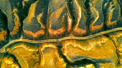 Aerial View of Car on Dirt Road. Drone shot of a white car driving on a winding dirt road through rugged golden hills and valleys at sunset. © Iryna