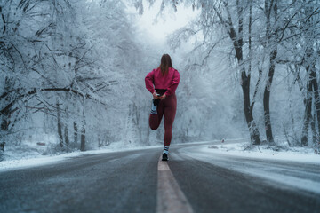 Young sportswoman stretching her legs in winter sportswear, preparing for a run along a snowy road,...