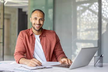 Obraz premium Portrait of a young successful businessman financier at his workplace inside the office, the employee is smiling and looking at the camera, writing down data on a paper report document
