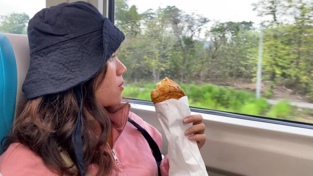 Beautiful young asian woman wearing black bucket hat eating croissant bread while looking into train window in train car during sunny day. Concept for Solo traveling, vacation, holiday, journey, trip.