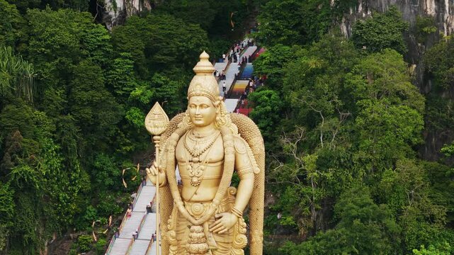 Large golden murugan statue towering near colorful limestone staircase ascending to sacred hindu temple entrance in batu caves, malaysia