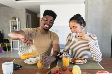 A smiling couple enjoying breakfast together at home, with the man pouring orange juice into a glass and the woman sipping her drink while looking at her phone, seated at a wooden table
