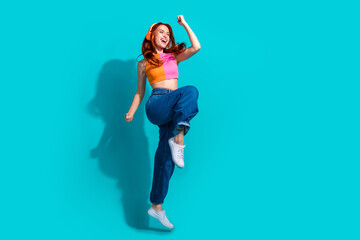 Vibrant young woman with red hair in summer casual outfit jumps joyfully against a bright teal...