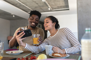Happy couple taking a selfie while enjoying breakfast together at home, capturing a joyful and candid moment in a modern, cozy dining area