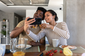Smiling couple sitting at a table, taking a selfie together on a smartphone, sharing a joyful moment in a modern home environment with a casual vibe