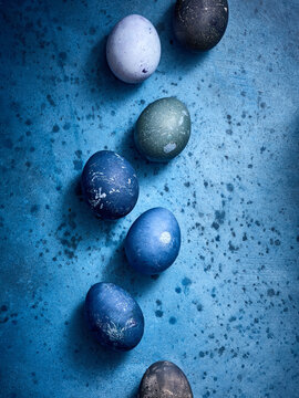 A variety of beautifully dyed eggs in shades of blue, green, and black are arranged in a straight line on a textured blue background. Perfect for spring festivities