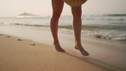 Barefoot woman walks on sunny beach, waves wash over her feet. Female holds hat, enjoys seaside stroll, experiences seaside freedom. Leisure, vacation vibes at coastal retreat in summer.