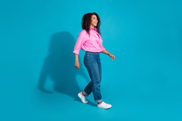 Charming young woman in casual pink shirt smiling and walking confidently against a vibrant blue...
