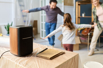 Notepad with sketches and portable stereo speaker standing on wooden piece of furniture against happy family dancing