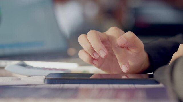 A person is using a cell phone with a finger on the screen. The phone is on a table with a laptop and a stack of papers. The scene suggests that the person is multitasking