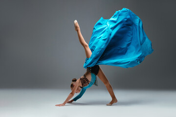 Graceful ballerina dancing on grey studio background. Cheerleader dancing with flying blue cloth fabric. Dancer in sport leotard doing Split. Child, artist, movement, action and motion concept