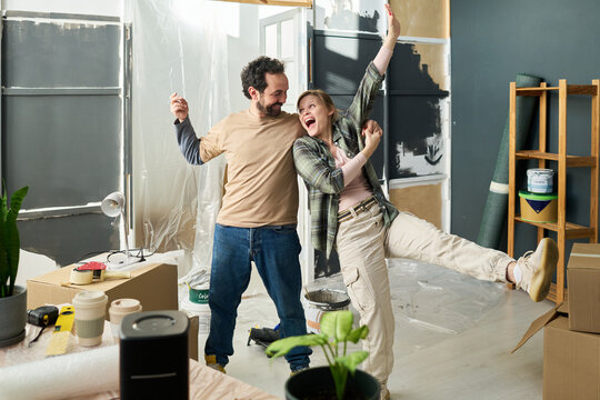 Young excited woman looking at her smiling husband and holding him by hand while dancing during renovation of living room