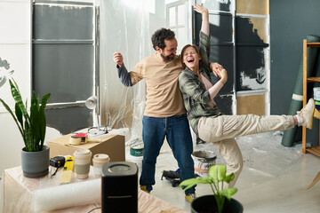 Young excited couple in casual apparel holding by hands and dancing at break during renovation of domestic room