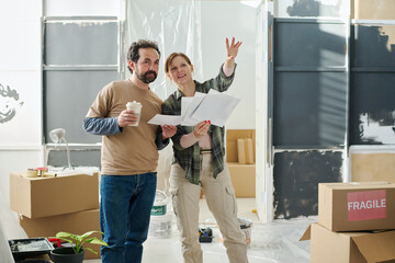 Young woman with several interior sketches pointing at wall of living room while explaining her...