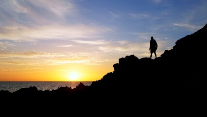 Fototapeta premium Silhouette of a solo traveler standing on volcanic rocks, enjoying a beautiful sunset over the Atlantic Ocean in the Canary Islands, Spain, feeling a sense of freedom and adventure