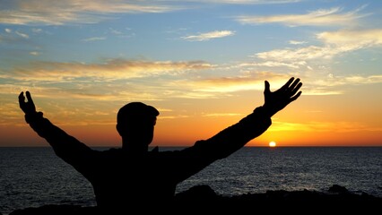 Fototapeta premium Silhouette of a young traveler raising arms in front of a beautiful sunset over the ocean in Canary Islands, celebrating freedom and enjoying the moment of solitude