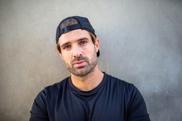 Man in black t-shirt and cap looking at the camera against a plain wall