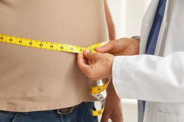 Nutritionist measuring overweight man's waist with tape in clinic, closeup