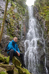 Tourist attraction of Germany - falls of Burgbach Waterfall near Schapbach, Black Forest, Baden-Wurttemberg, Germany. Man hiker in blue jacket standing on stone and looks at flow of falling water